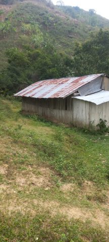 Wooden school building with a corrugated metal roof on a grassy hillside below forest