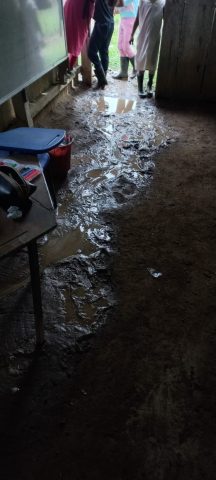 Mud and standing water across a dirt classroom floor beside a desk, students in rubber boots at the door