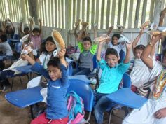 Smiling students at blue desk-chairs raise snacks in the air inside a wooden-slat rural classroom