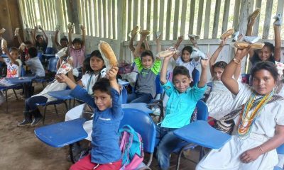 Smiling students at blue desk-chairs raise snacks in the air inside a wooden-slat rural classroom