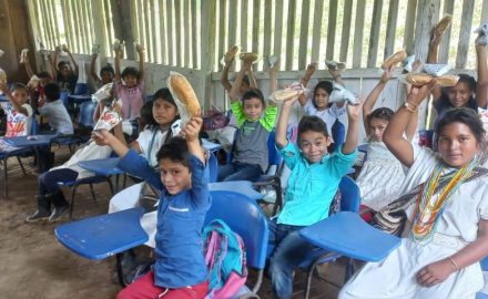 Smiling students at blue desk-chairs raise snacks in the air inside a wooden-slat rural classroom