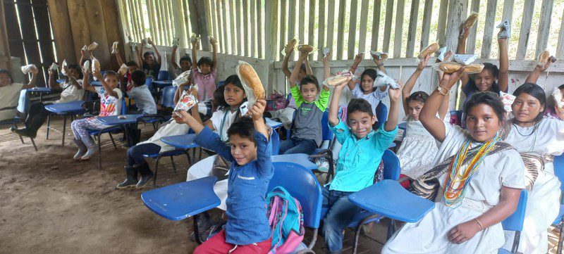 Students seated at blue desk-chairs hold snacks overhead in an open-sided wooden classroom