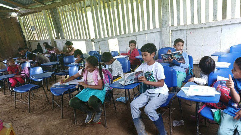 Students read at blue plastic desk-chairs set on a dirt floor inside the school