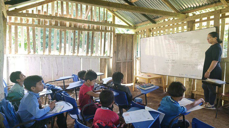 Teacher stands at a whiteboard while students work at desk-chairs inside a wooden-slat classroom
