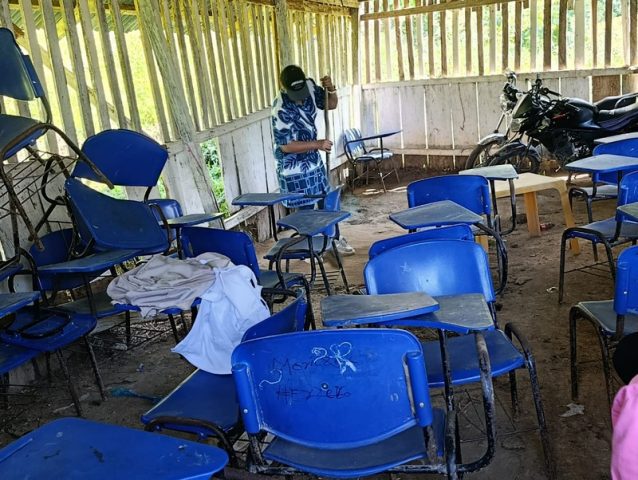 Person sweeps a dirt floor among worn blue desk-chairs, a motorcycle parked inside the classroom