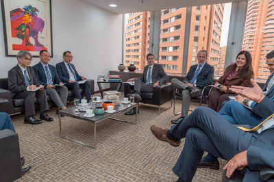 Officials sit on sofas and armchairs around a coffee table in an office meeting room with tall brick buildings outside.