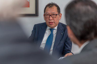 A man in glasses, a navy suit and a blue tie, wearing a Mazda lapel pin, speaks during a meeting.