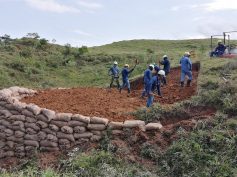 Workers in hard hats dig at a hillside site edged with a sandbag wall.