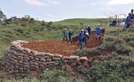 Workers in hard hats dig at a hillside site edged with a sandbag wall.