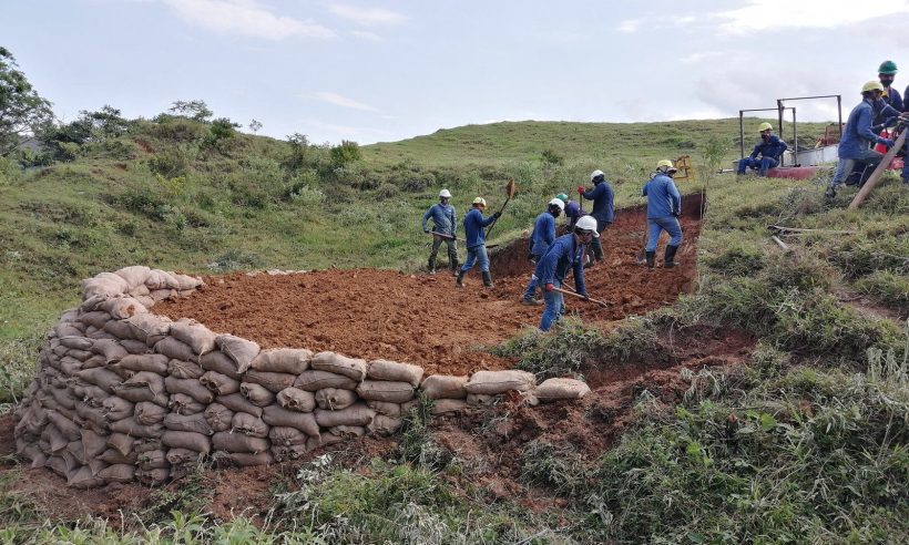 Workers in hard hats dig at a hillside site edged with a sandbag wall.