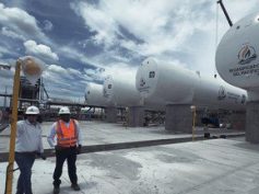 Two workers in hard hats walk past LNG storage tanks bearing the Regasificadora del Pacífico logo in El Vínculo, Buga