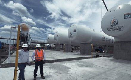 Two workers in hard hats walk past LNG storage tanks bearing the Regasificadora del Pacífico logo in El Vínculo, Buga