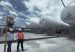 Two workers in hard hats walk past LNG storage tanks bearing the Regasificadora del Pacífico logo in El Vínculo, Buga