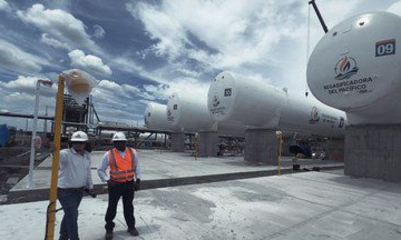 Two workers in hard hats walk past LNG storage tanks bearing the Regasificadora del Pacífico logo in El Vínculo, Buga